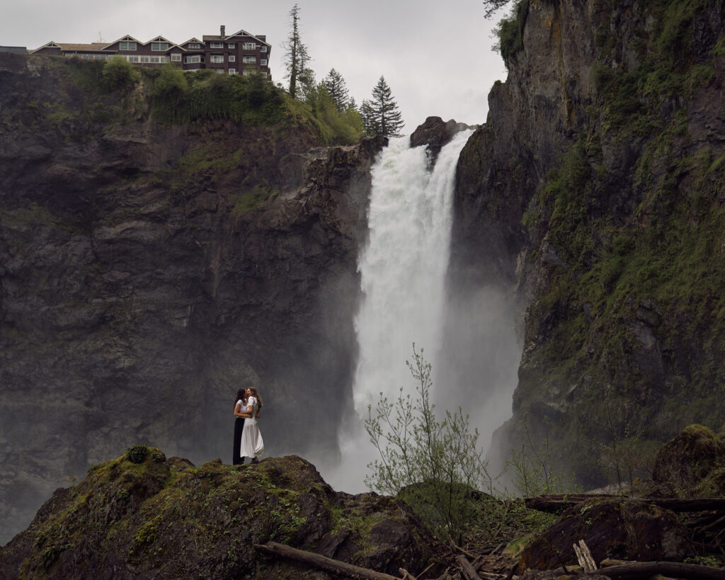 two brides hug in front of a waterfall