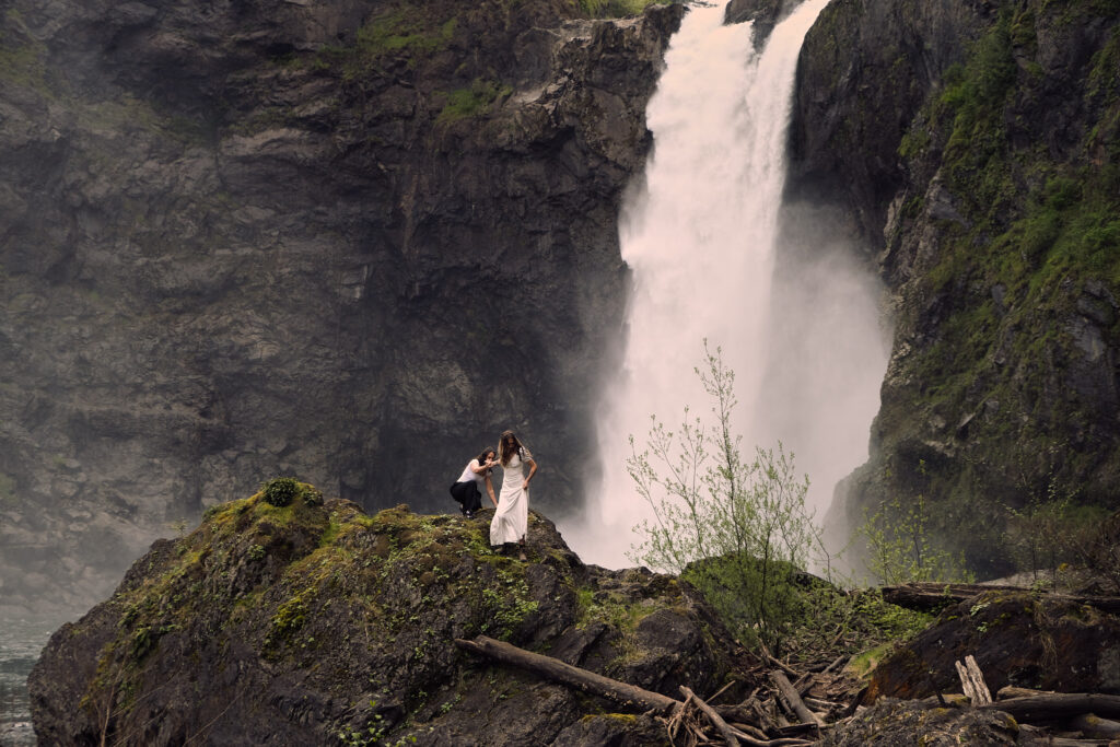 Two brides help each other over a rock in front of Snoqualmie Falls