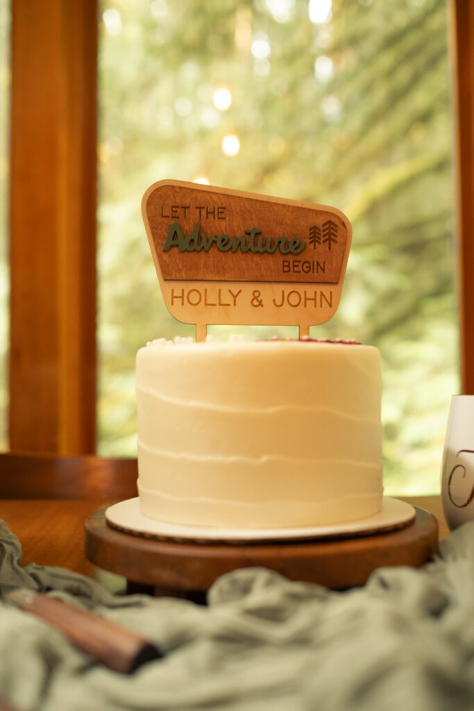 a small white frosted cake with a sign in a national park style reads "Let the Adventure Begin"