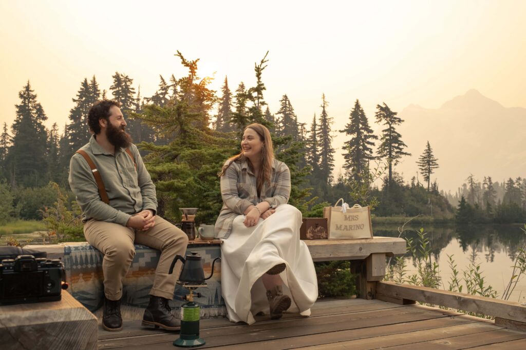 a bride and groom sit on a dock at sunrise in a smokey haze